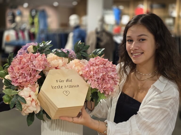 Junge Frau präsentiert eine Muttertags-Holzbox mit der Aufschrift „Für die beste Mama“ zwischen rosa Hortensien im Echter Modehaus in Murnau als liebevolle Geschenkidee zum Muttertag.