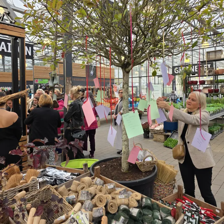 Frau hängt bunte Wunschkarten an einen "Wunsch"-Baum im Kiefl Gartencenter in Gauting, umgeben von Besucherinnen.