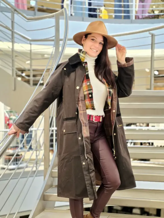 Andrea aus Ohlstadt präsentiert einen eleganten Herbst-Winter-Look mit langem Barbour-Mantel, kariertem Innenfutter, Rollkragenpullover, brauner Hose und beigem Hut auf einer Treppe im Modehaus.