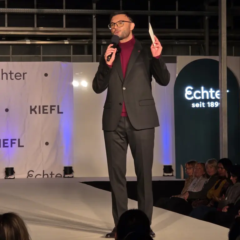 Ein Mann im schwarzen Anzug spricht mit Mikrofon auf der Bühne der Echter Modenschau. Das ist der Moderator Egzon Maliqi.