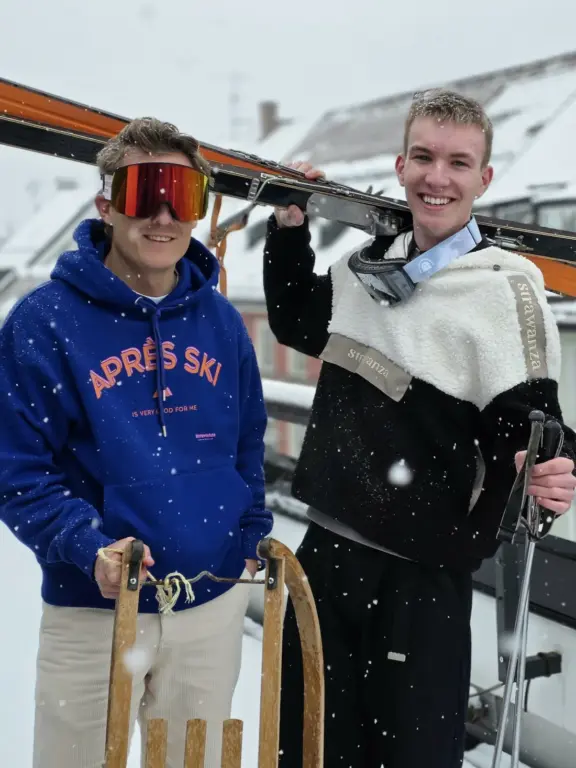 Fabia und Jakob aus dem Oberland posieren im Schnee mit Retro-Ski und Schlitten in farbigen Strawanza-Looks mit Apré-Ski-Stimmung.