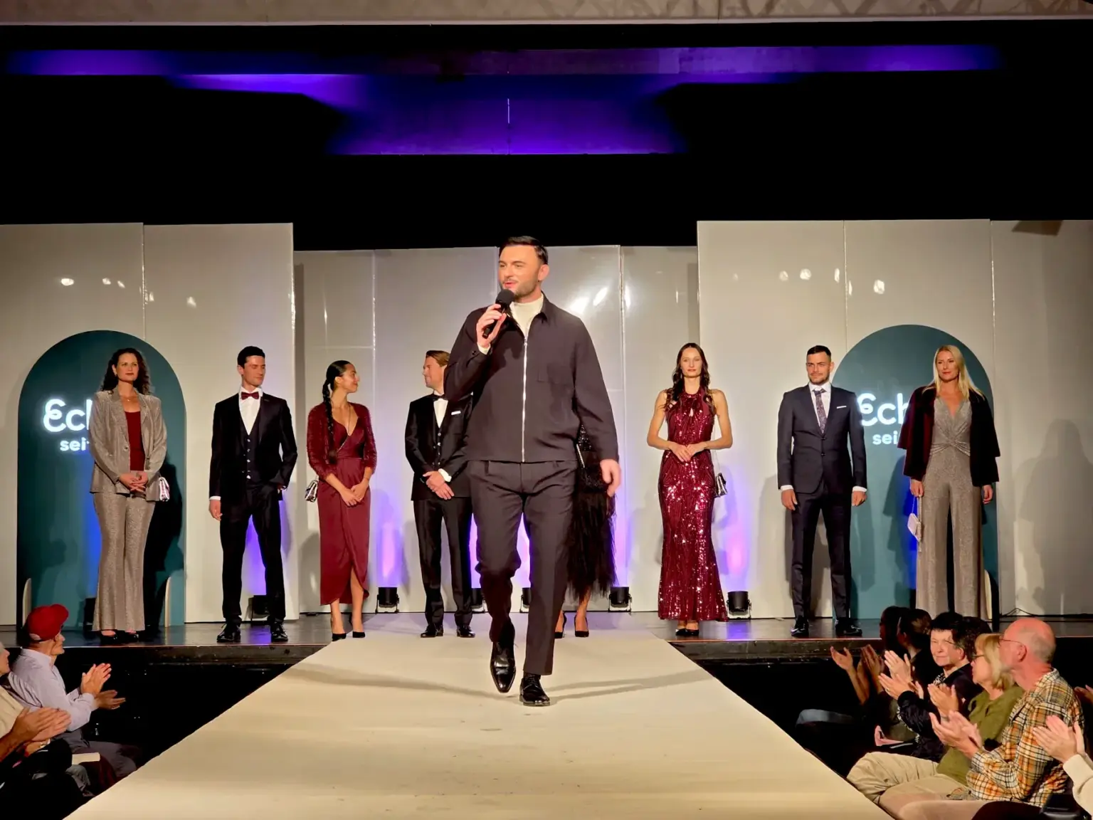 Das Finale der Großen Echter Show zur Kunstnacht am 2.10.25 in KTM Murnau. Der Moderator und die Models stehen festlich und freudig auf der Bühne und Zuschauer klatschen.