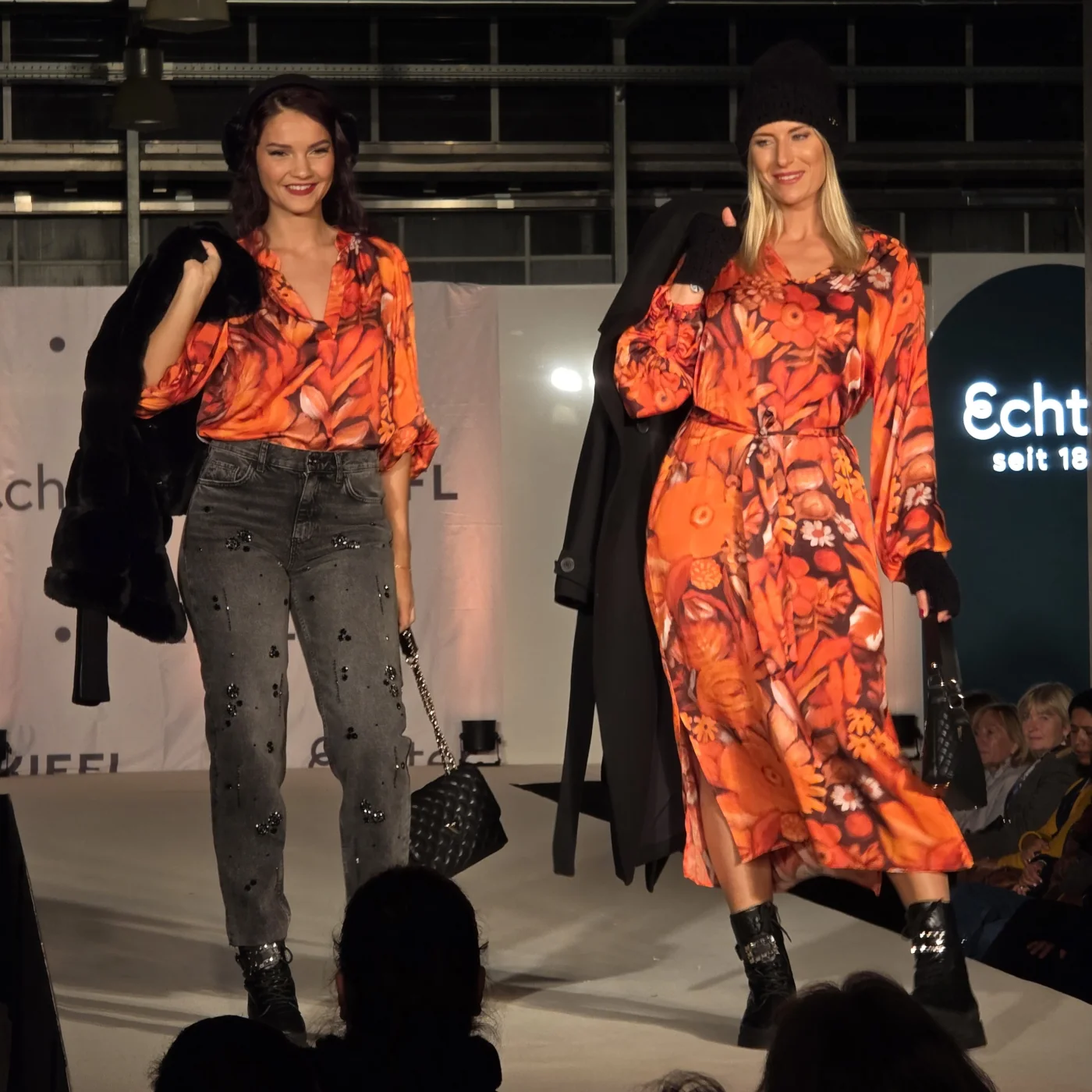 Zwei Models tragen orangefarbene Blusen mit floralen Mustern und kombinieren sie mit Jeans und Kleid. Sie stehen auf der Kiefl-Bühne im Gartencenter bei der Großen Echter Modenschau.