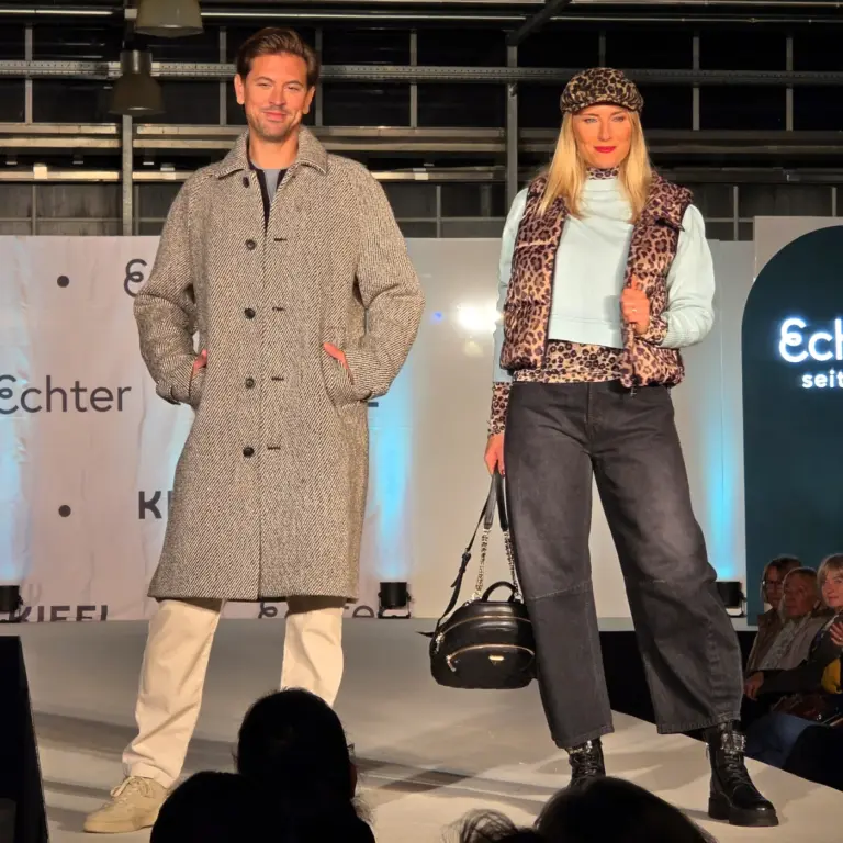 Zwei Models präsentieren Outfits mit Mantel, Jeans, Leo-Weste und hellblauem Pullover auf der Kiefl-Bühne im Gartencenter in Gauting