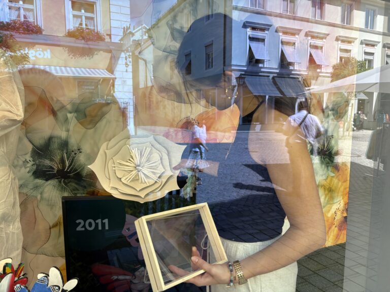 Tabea von Echter beim Arrangieren von Deko oder Bildern im Schaufenster von Echter Mode in Murnau, im Hintergrund florale Gestaltungselemente und Reflexion der Fußgängerzone.