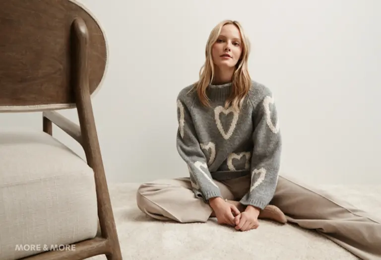 Model trägt einen grauen Strickpullover mit cremefarbenem Herzmuster und beigefarbener Hose, sitzend auf Teppichboden vor neutralem Hintergrund – Kollektion von MORE & MORE