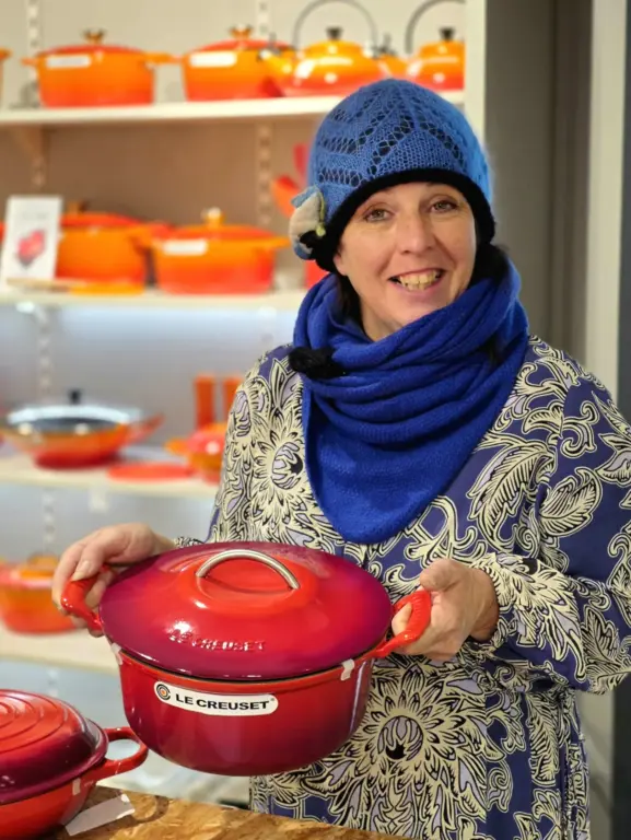 Rosa aus Uffing präsentiert einen roten Le Creuset Bräter vor einem Regal voller farbiger Gusseisen-Kochgeschirre.