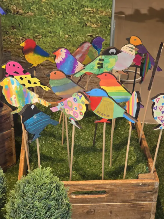 Viele farbenfroh bemalte Holzspatzen auf Holzstäben in einer Holzkiste.