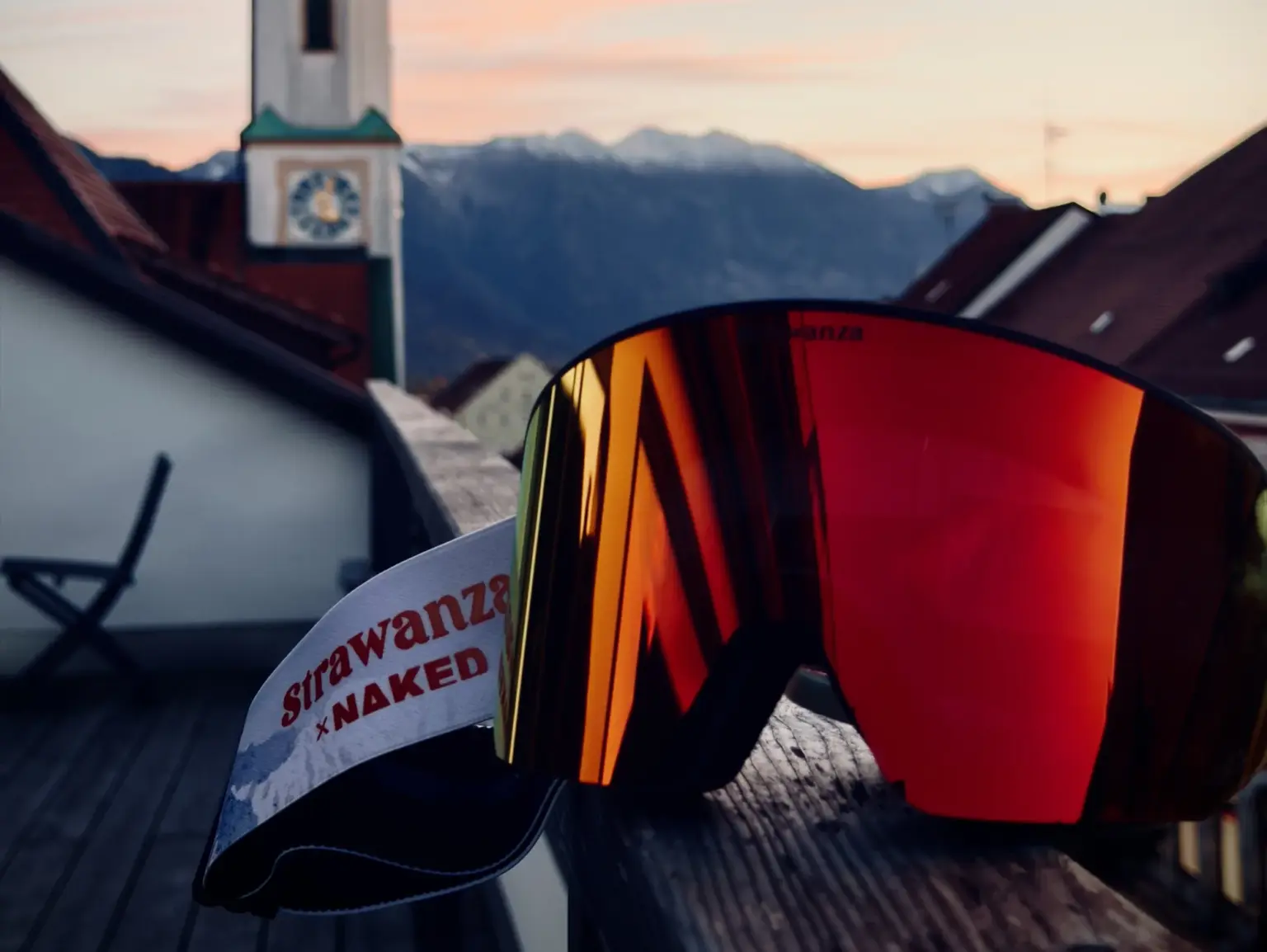 Skibrille STRAWANZA mit rot verspiegelter Scheibe auf Holzgeländer; im Hintergrund Murnauer Kirchturm und Alpen im Abendlicht.