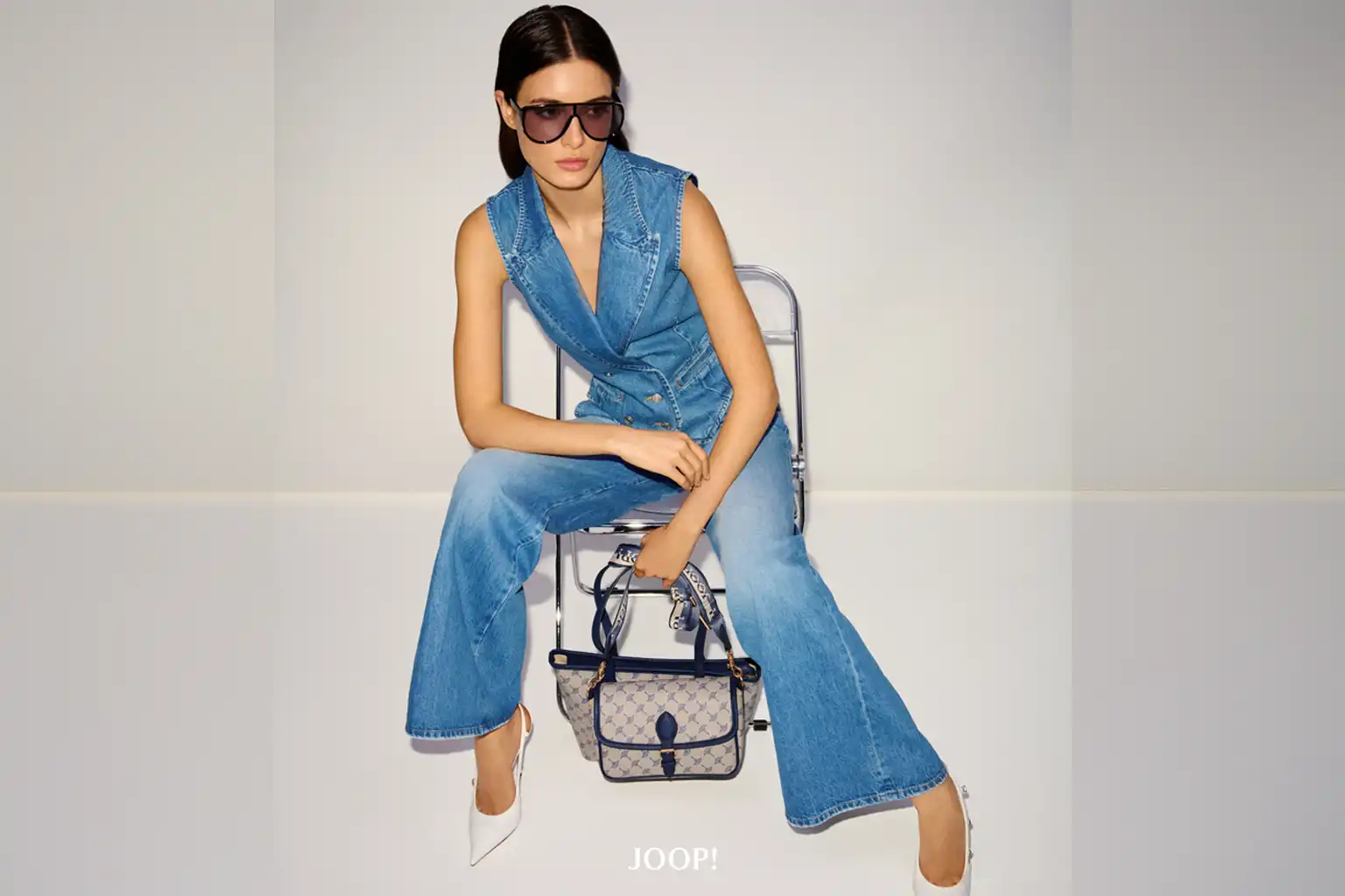Model in ärmellosem Jeansanzug von JOOP! mit weiter Hose, weißem Schuh und Designer-Handtasche – fotografiert im Studio auf Stuhl sitzend.