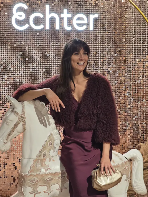 Lächelnde Frau in einem bordeauxfarbenen Satinkleid mit flauschiger Kurzjacke und goldener Clutch neben einem weißen Dekopferd. Im Hintergrund eine glitzernde Wand mit leuchtendem Echter-Schriftzug im Modehaus Echter in Weilheim.