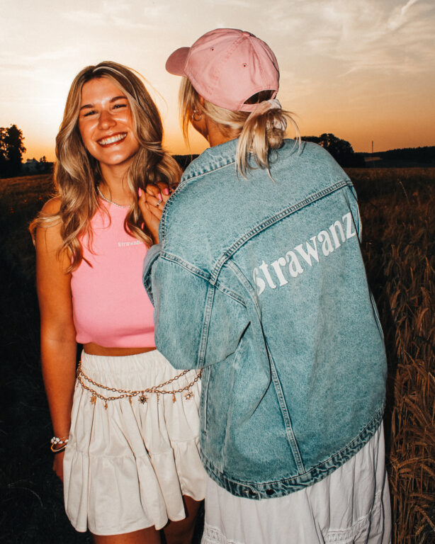 Zwei junge Frauen in Strawanza-Outfits beim Sonnenuntergang, eine mit Jeansjacke und Cap, die andere in pinkem Top, beide lachen.